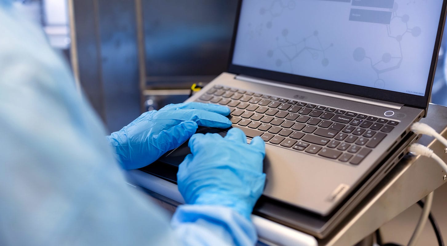 A person wearing blue medical gloves types on a laptop keyboard, with a chemical structure displayed on the screen.